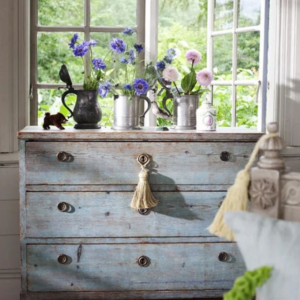 Distressed blue farmhouse dresser with tassel pulls, vintage pitchers, and fresh flowers by window