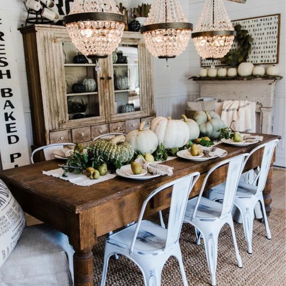 Rustic farmhouse dining table with white metal chairs, crystal chandeliers, and autumn pumpkin centerpiece