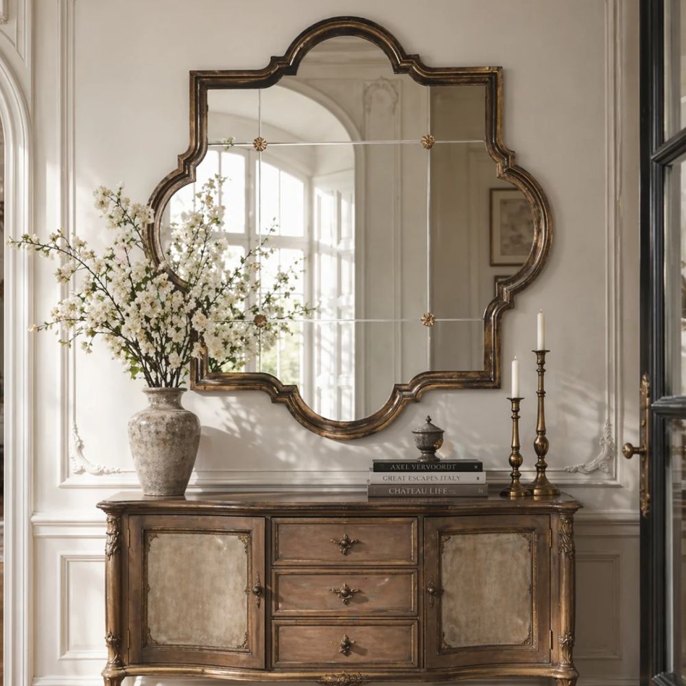 An ornate arched mirror with a distressed bronze-gold frame hangs above a matching wooden console table in a classical interior setting with a vase of white blossoms and candlesticks.