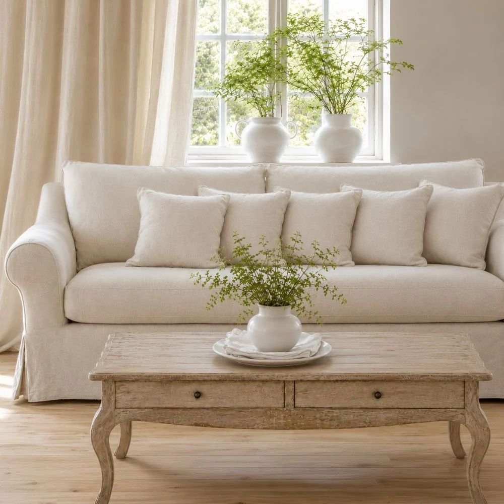 Natural French country living room with cream slipcover sofa, rustic carved wood coffee table, linen curtains, and fresh greenery