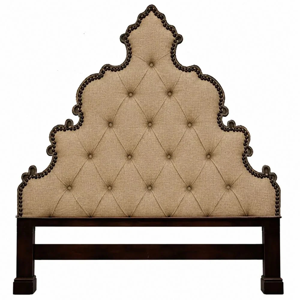 An ornate, high-garland headboard with a beige tufted linen fabric and dark brown carved wood trim with nailhead detailing, supported by a dark brown wooden frame.